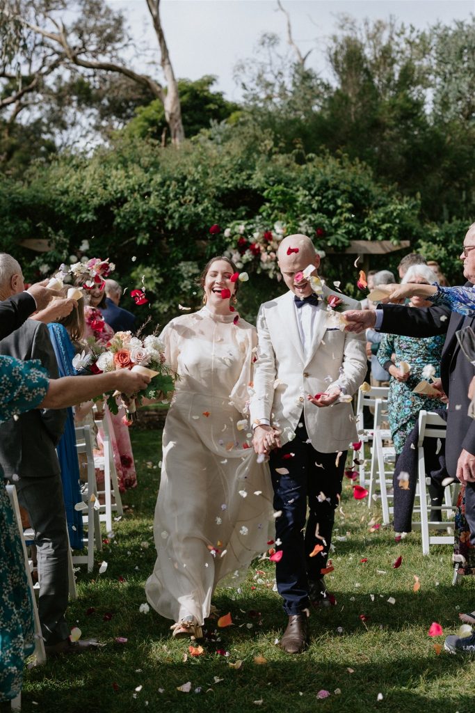 A bride and groom walk down an aisle with flower petal confetti being thrown, they are in a garden with smiling guests