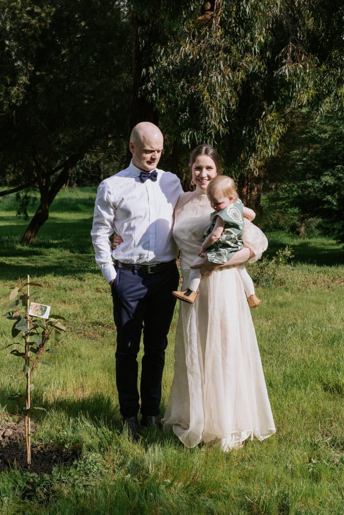 A bride and groom wearing ethically made wedding dress and suit, stand next to a tree they planted on their wedding day