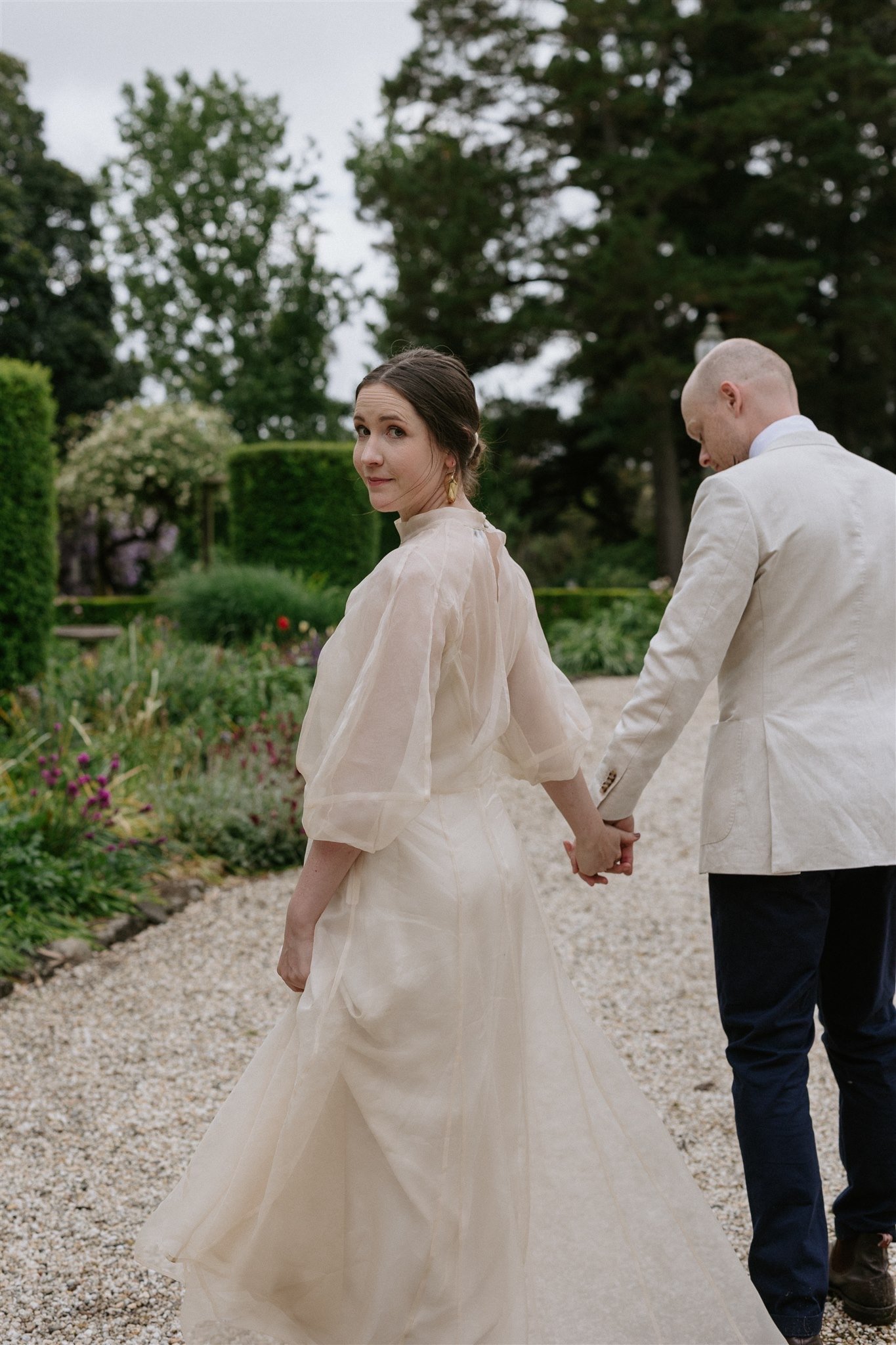 A bride wearing an ethically made wedding gown looks back at the camera while walking through an elegant garden