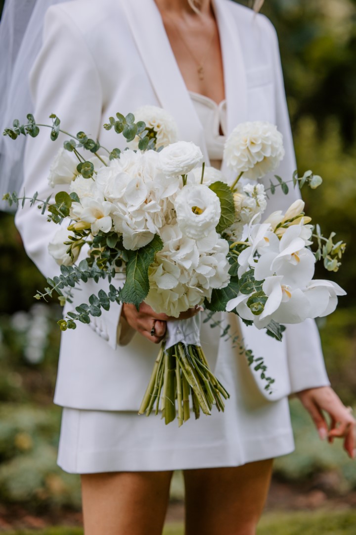 A bride wearing a veil and suit jacket holds a bouquet of white flowers. She stands in the garden at Highwic in Newmarket - Central Auckland.